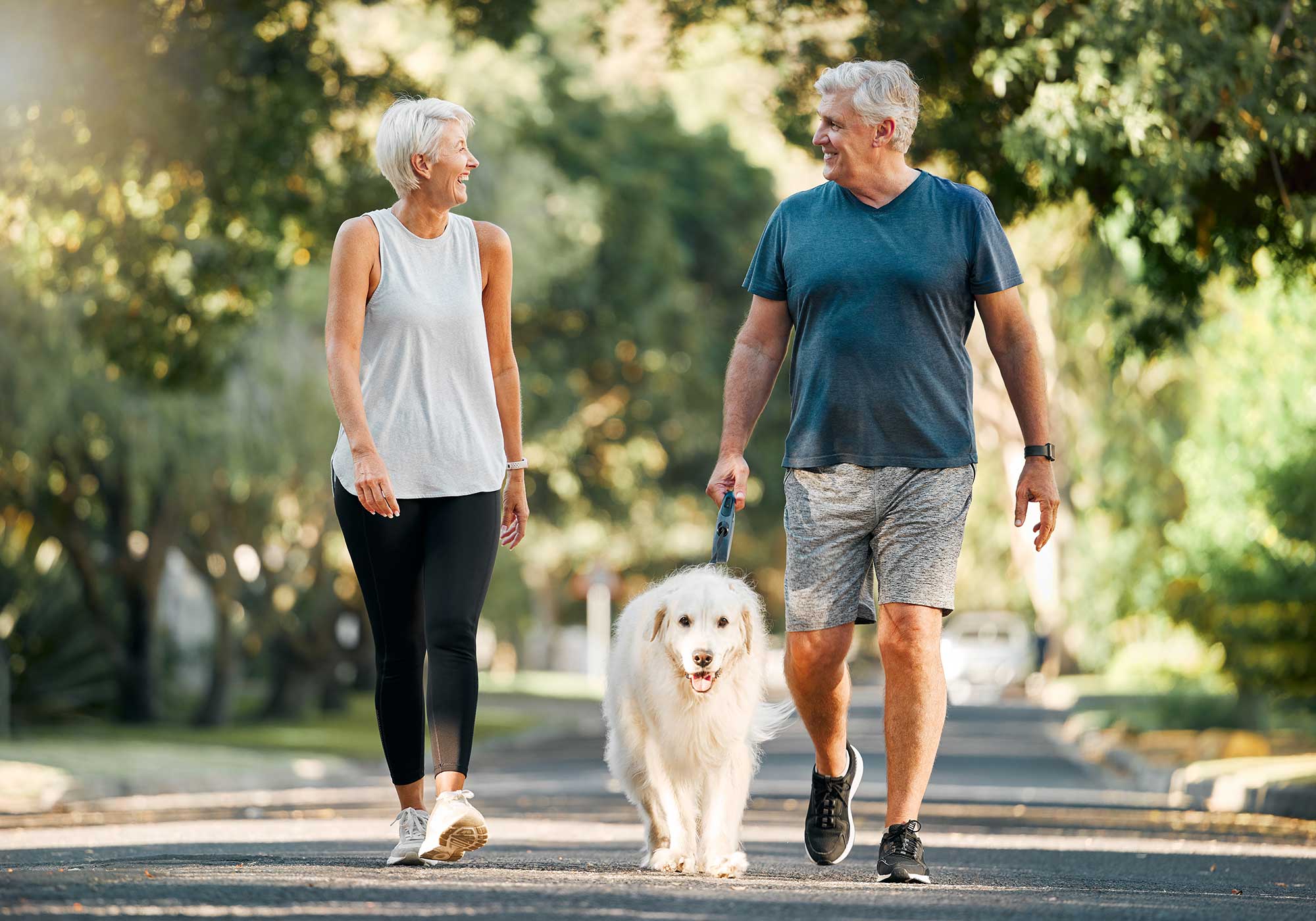 Couple smiling looking at each other while walking their dog on a sunny day. 