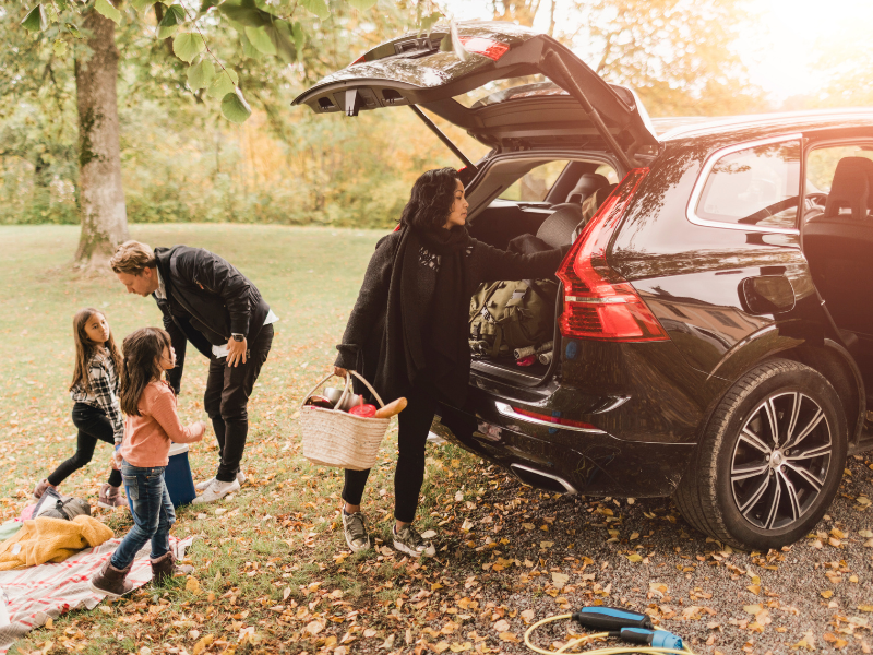Mother unloading bags from the back of an SUV as children play nearby in a park with their father.