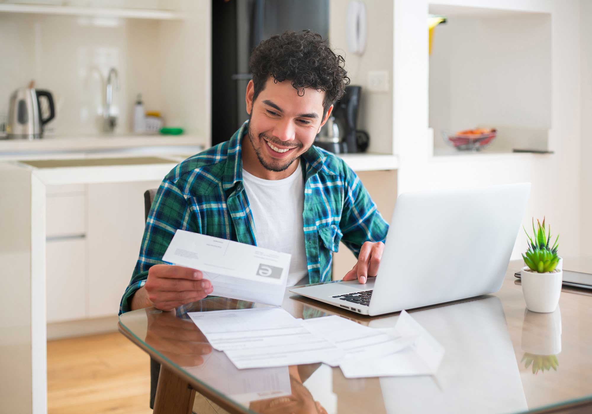 A man sitting in his kitchen smiling while looking at his bank statement. 