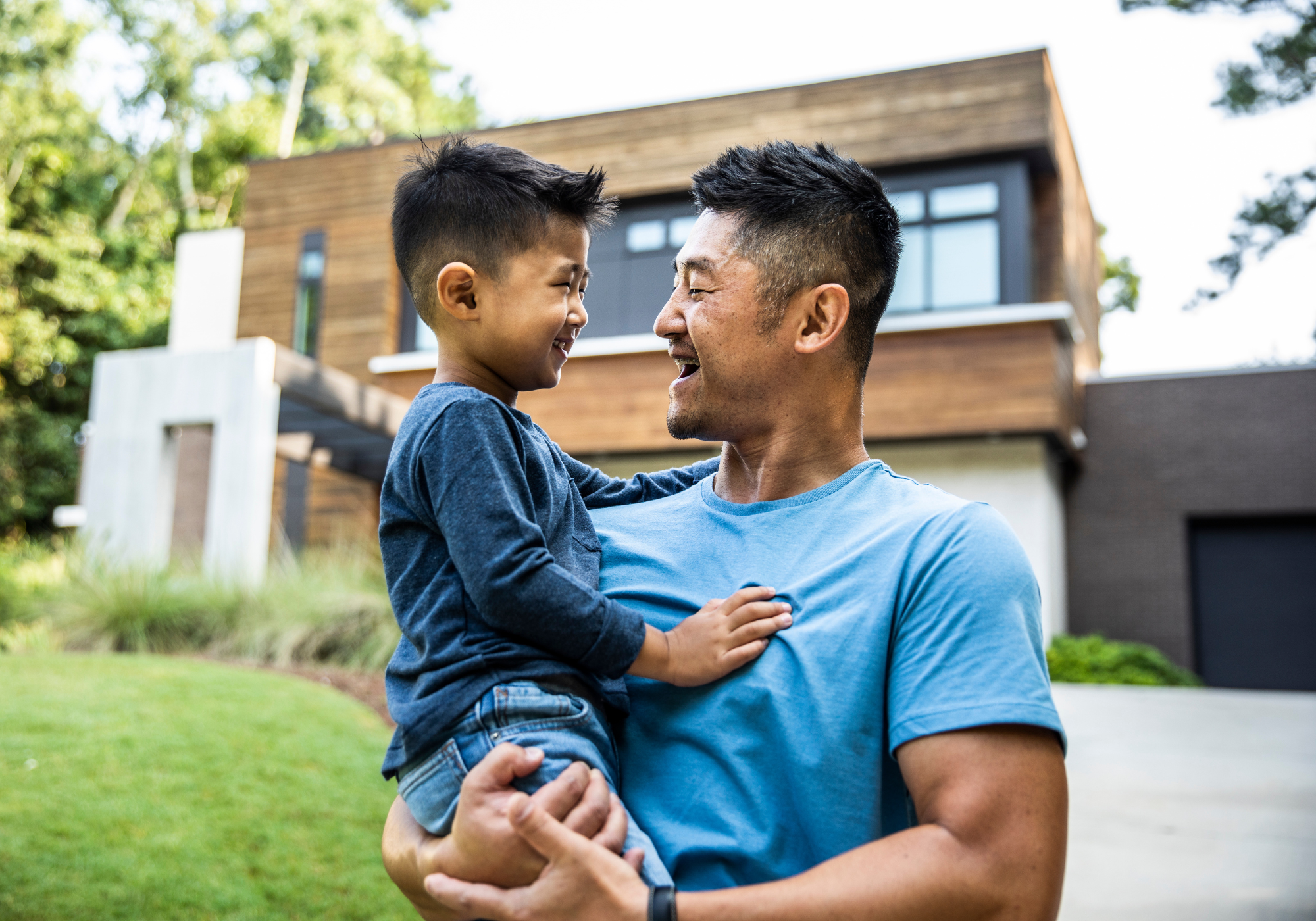 Father holding young son while smiling and standing outside of home.