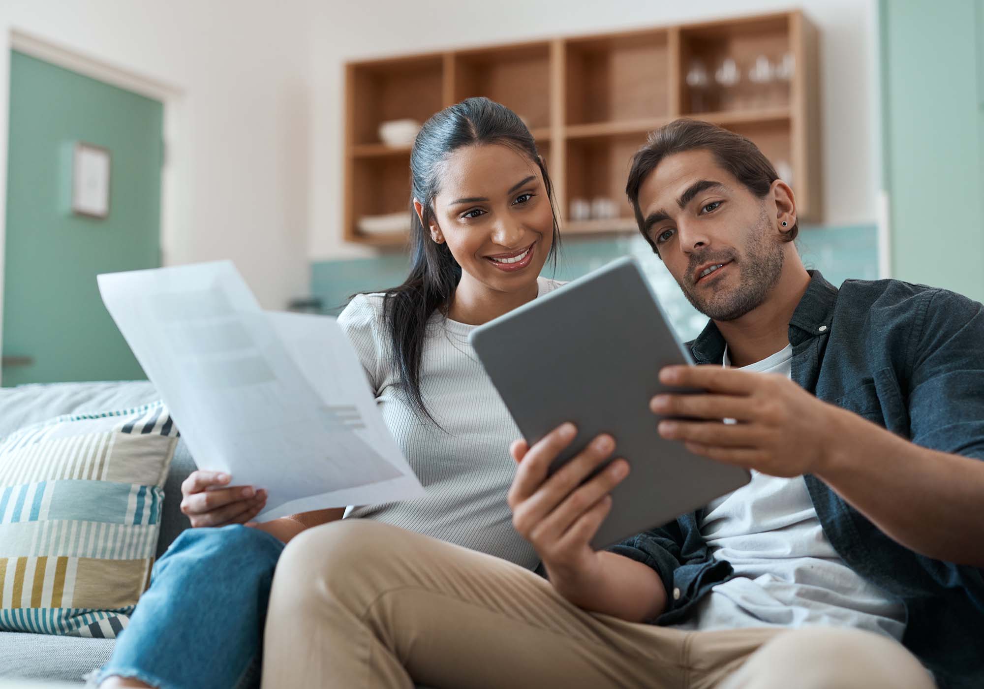 Couple sitting on the sofa looking at their bank statements on a laptop and paper. 