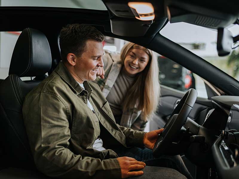 A man sitting in the driver's seat of a car with the door open and a woman looking in smiling. 