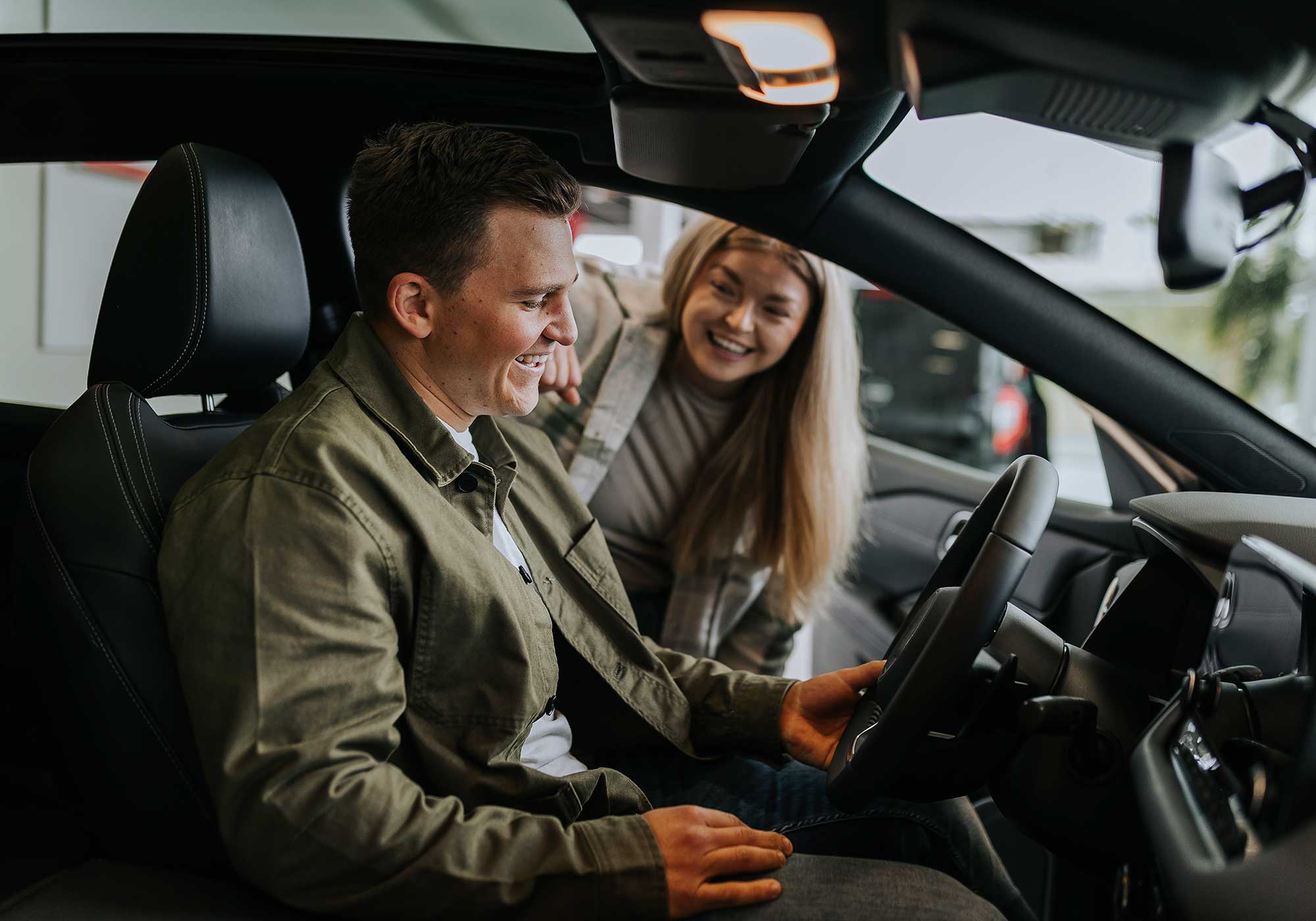 Man sitting in a car holding the wheel, while a woman leans in smiling. 