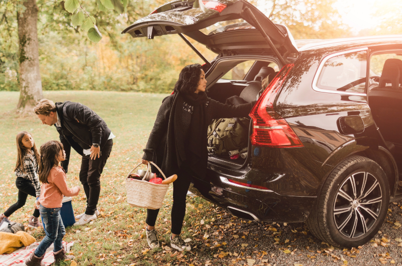 Mother unloading bags from the back of an SUV as children play nearby in a park with their father.